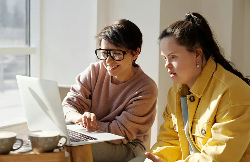 Zwei Personen sitzen an einem Tisch und schauen auf einen Laptop; eine Person trägt eine Brille, die andere hat Down-Syndrom. Auf dem Tisch stehen zwei Tassen.