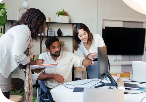 Drei Personen in modernem Büro schauen auf einen Monitor: eine steht und schreibt in Notizblock, eine zeigt auf den Bildschirm, eine lehnt sich zum Bildschirm.
