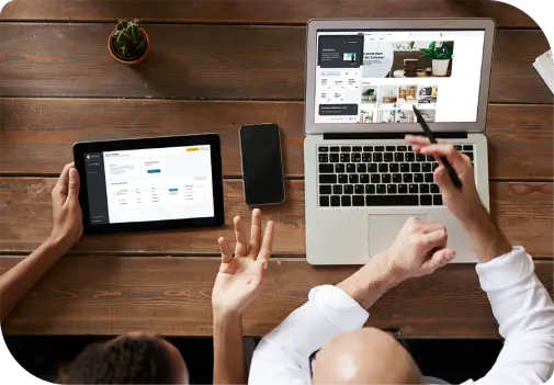 Zwei Personen an Holztisch: eine zeigt mit Stift auf Laptop, die andere hält Tablet; auf Laptop Website mit danova Assistant, auf Tablet Dashboard sichtbar.